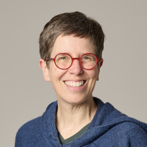Anita Verna Crofts headshot, smiling warmly with red rimmed glasses and a blue sweatshirt